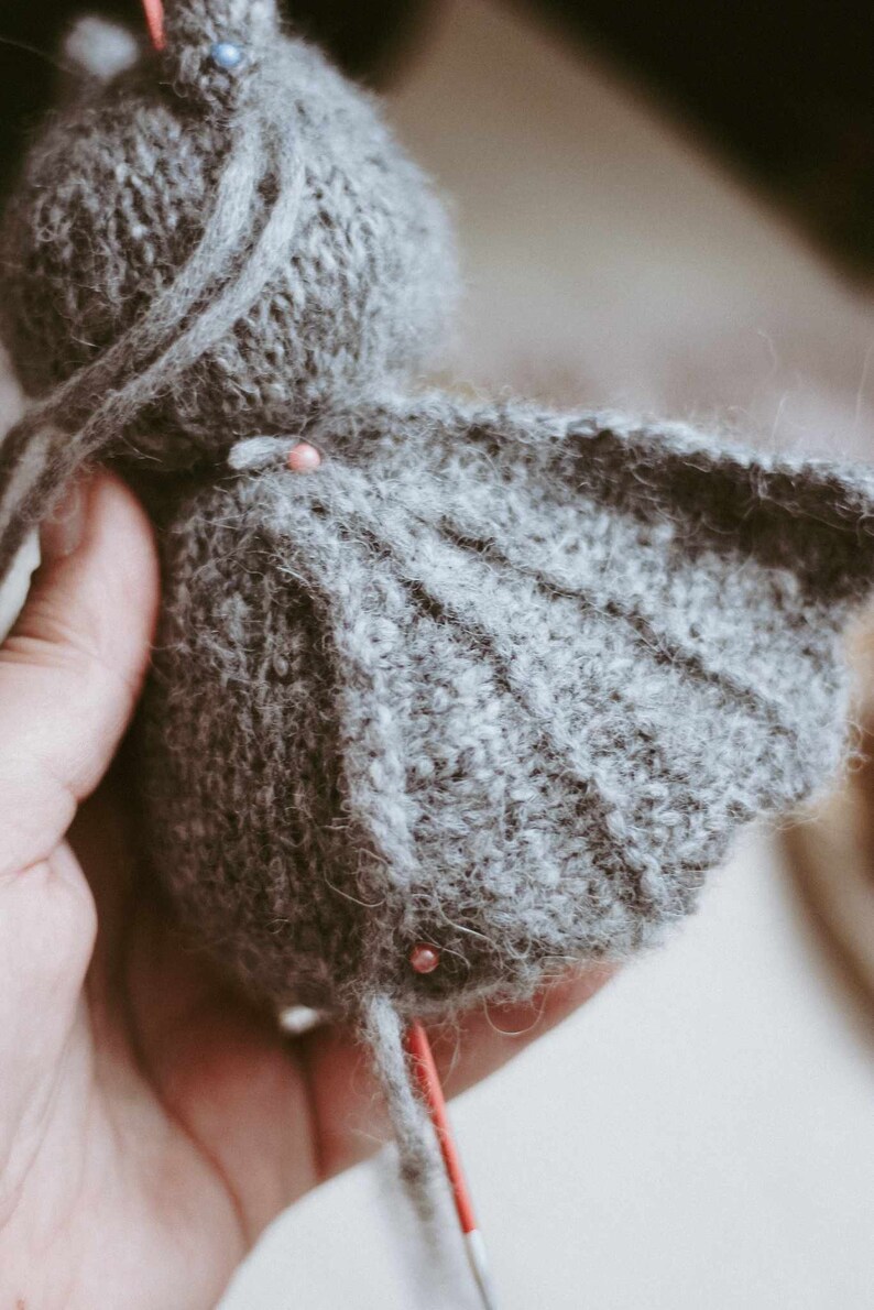 May include: A gray knitted stuffed animal with a long tail and wings. The animal is being sewn together with red pins.