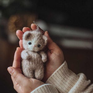 May include: A small, gray and white knitted cat toy held in cupped hands. The cat toy has a white face with black eyes and a pink nose.