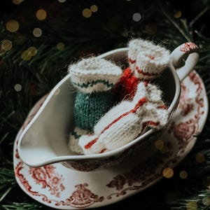 May include: A white gravy boat with a red floral pattern holds three miniature knitted Christmas stockings. One stocking is green and white, another is red and white, and the third is white with red stripes. The scene is set against a backdrop of evergreen branches.
