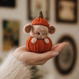 Pode incluir: Uma pequena figura de rato tricotada à mão com pelo bege claro, orelhas redondas e um nariz minúsculo cor-de-rosa. O rato usa um gorro laranja canelado e está aninhado dentro de uma base tricotada laranja, redonda e texturizada em forma de abóbora.
