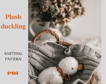 May include: A white plush duckling toy with brown feet and a red ribbon is nestled in a wicker basket lined with a gray knitted blanket. The text "Plush duckling" is in orange text at the top of the image. The text "Knitting Pattern" is in orange text below the image. The text "PDF" is in orange text at the bottom of the image.