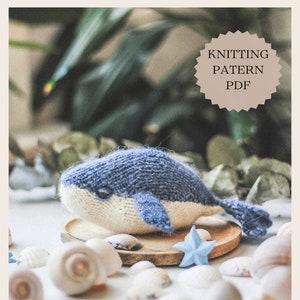 May include: Knitted whale amigurumi toy in blue and white, sitting on a wooden surface with seashells and a blue starfish.  The image includes the text "Knitting Patern PDF".