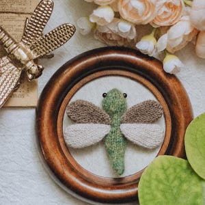 May include: A knitted green dragonfly with brown and cream wings, mounted inside a circular dark wood frame. The dragonfly features two small black bead eyes and a textured yarn body, displayed alongside a gold metal dragonfly ornament.