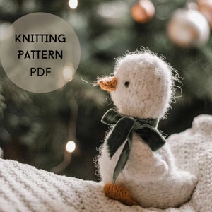 Könnte beinhalten: Strickmuster PDF für eine weiße, flauschige Ente mit einem grünen Samtband um den Hals. Die Ente sitzt auf einer weißen Strickdecke.