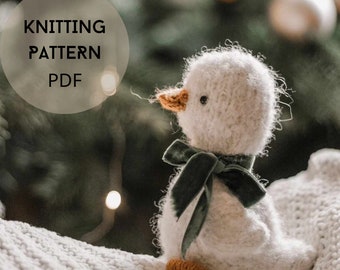 May include: Knitting pattern PDF for a white fluffy duck with a green velvet ribbon around its neck. The duck is sitting on a white knitted blanket.