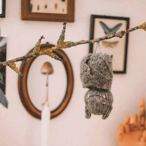 May include: A gray knitted bat hanging from a branch with a brown oval frame in the background.