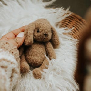 May include: A small, handmade brown bunny plush toy with floppy ears and a stitched face. The bunny is resting on a fluffy white surface. The toy is held by a person wearing a beige sweater.