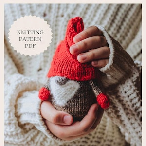 May include: A knitted gnome toy with a red hat, white beard, and brown body. The gnome is being held by a person's hands.