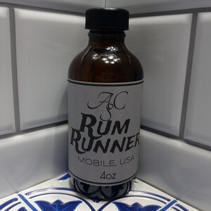 May include: A brown glass bottle with a black cap and a gray label. The label reads "AC Rum Runner MOBILE, USA 4oz". The bottle is sitting on a blue and white patterned surface.