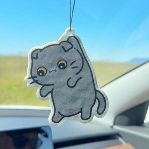 K&ouml;nnte beinhalten: Ein grau geformter Lufterfrischer in Katzenform, der an einer Schnur in einem Auto h&auml;ngt. Die Cartoon-Katze hat gro&szlig;e gelbe Augen und einen wei&szlig;en Umriss. Der Hintergrund zeigt einen blauen Himmel und ein gr&uuml;nes Feld.