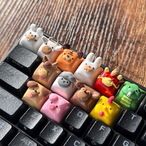 May include: A keyboard with custom keycaps featuring 12 different animal designs. The keycaps are made of polymer clay and are painted in various colors. The keycaps are arranged in a row across the keyboard, starting with a white rabbit and ending with a green snake. The text "small hoogin studios" is visible in the upper right corner of the image.