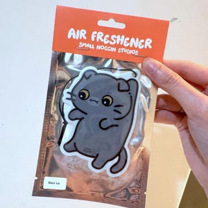 K&ouml;nnte beinhalten: Lufterfrischer in einer silbernen Verpackung mit einem Cartoon-Katzen-Design. Der orangefarbene Header lautet "AIR FRESHENER" und "SMALL NOGGIN STUDIOS". In der unteren linken Ecke steht "Black ice".