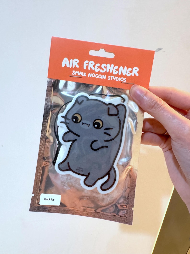 K&ouml;nnte beinhalten: Lufterfrischer in einer silbernen Verpackung mit einem Cartoon-Katzen-Design. Der orangefarbene Header lautet "AIR FRESHENER" und "SMALL NOGGIN STUDIOS". In der unteren linken Ecke steht "Black ice".