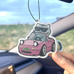 May include: A car air freshener featuring a cartoon cat wearing sunglasses driving a pink convertible. The car has the text "SPALL NOCON" on the license plate. The air freshener hangs from a black string.