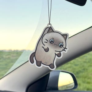 Könnte beinhalten: Ein Auto-Lufterfrischer mit einem Cartoon-Katzen-Design. Die Katze ist hellbraun mit dunkelbraunen Akzenten und einem schwarzen Umriss. Er hängt an einer schwarzen Schnur, sichtbar im Inneren eines Autos.