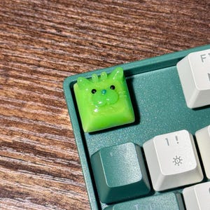 May include: A bright lime green artisan keycap shaped like a cat face with small ears, two black eyes, and a tiny blue nose, installed on a dark green mechanical keyboard next to white keys featuring a sun icon and the number one with an exclamation mark.