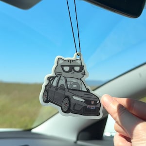 May include: A car air freshener featuring a cartoon cat wearing sunglasses atop a gray car. The air freshener is hanging from a black string, with a blurred background of a blue sky and green field.