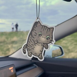 Könnte beinhalten: Ein hängender Auto-Lufterfrischer mit einem Cartoon-Katzen-Design. Die Katze ist grau mit dunkelgrauen Streifen, grünen Augen und einer weißen Umrandung. Der Lufterfrischer hängt an einer schwarzen Schnur in einem Auto.