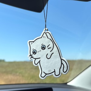 Könnte beinhalten: Ein weißer, katzenförmiger Lufterfrischer mit schwarzen Umrissen und Details, der an einer schwarzen Schnur hängt. Die Katze hat blaue Augen und eine verspielte Pose. Der Hintergrund zeigt einen blauen Himmel und eine ferne Landschaft.