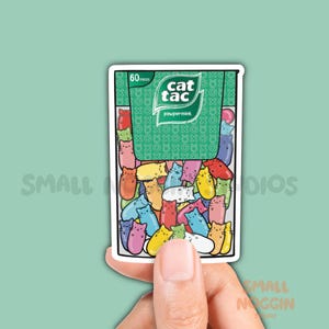 May include: A rectangular sticker featuring a green package labeled "cat tac" and "pawpermint" with "60 pieces" printed at the top. The lower portion of the package is transparent, revealing a collection of small, colorful, cat-shaped candies.