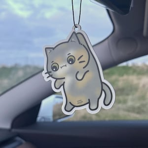Könnte beinhalten: Ein grau-gelber, katzenförmiger Lufterfrischer, der an einer schwarzen Schnur hängt. Die Cartoon-Katze hat große Augen und lächelt. Der Hintergrund zeigt einen Autoinnenraum und den Himmel.