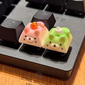 Pode incluir: Duas adoráveis teclas com forma de gato, uma rosa com um morango no topo e a outra verde com um redemoinho branco no topo. As teclas estão em um teclado preto.