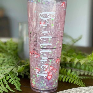 May include: A clear plastic tumbler with a pink and white glitter design. The tumbler has a white sticker with the name "Brittany" on it. The tumbler is filled with a pink liquid and has red strawberry pieces inside.