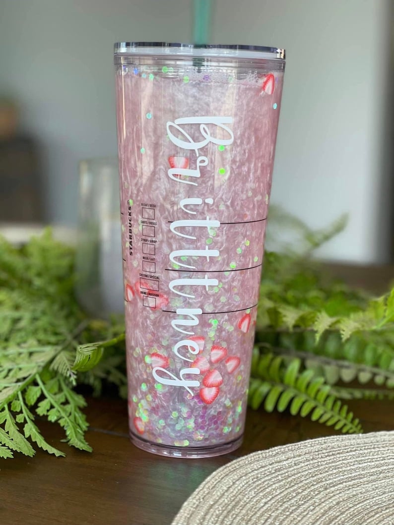 May include: A clear plastic tumbler with a pink and white glitter design. The tumbler has a white sticker with the name "Brittany" on it. The tumbler is filled with a pink liquid and has red strawberry pieces inside.