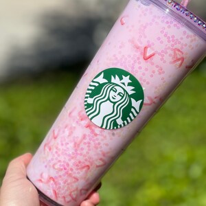 Peut inclure: Un gobelet r&eacute;utilisable rose avec un logo Starbucks vert et un motif de paillettes roses. Le gobelet a un couvercle transparent avec une paille rose.