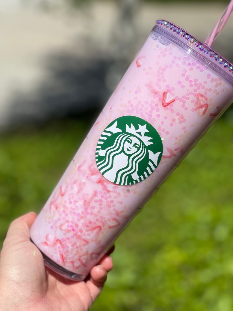 Peut inclure: Un gobelet r&eacute;utilisable rose avec un logo Starbucks vert et un motif de paillettes roses. Le gobelet a un couvercle transparent avec une paille rose.