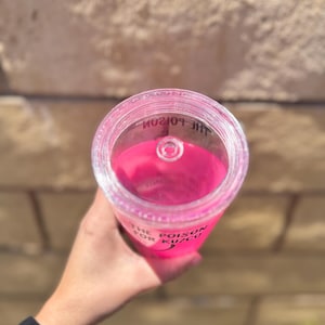 May include: A clear plastic tumbler filled with bright pink liquid. The text "THE POISON FOR KUZCO" is printed on the side. The tumbler has a clear lid with a small opening.