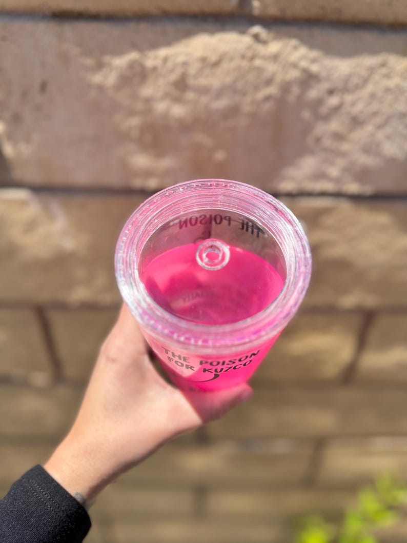 May include: A clear plastic tumbler filled with bright pink liquid. The text "THE POISON FOR KUZCO" is printed on the side. The tumbler has a clear lid with a small opening.