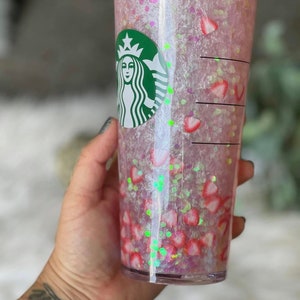 May include: A clear plastic tumbler with a pink and white glitter design. The tumbler has a green Starbucks logo on the side and is filled with pink and white glitter and strawberry slices.