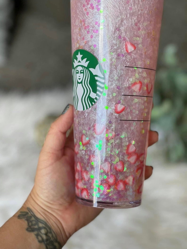 May include: A clear plastic tumbler with a pink and white glitter design. The tumbler has a green Starbucks logo on the side and is filled with pink and white glitter and strawberry slices.