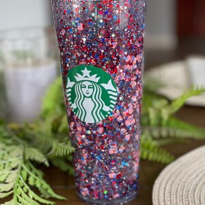 May include: A clear tumbler with a red, white and blue glitter design. The tumbler has the Starbucks logo on the front.