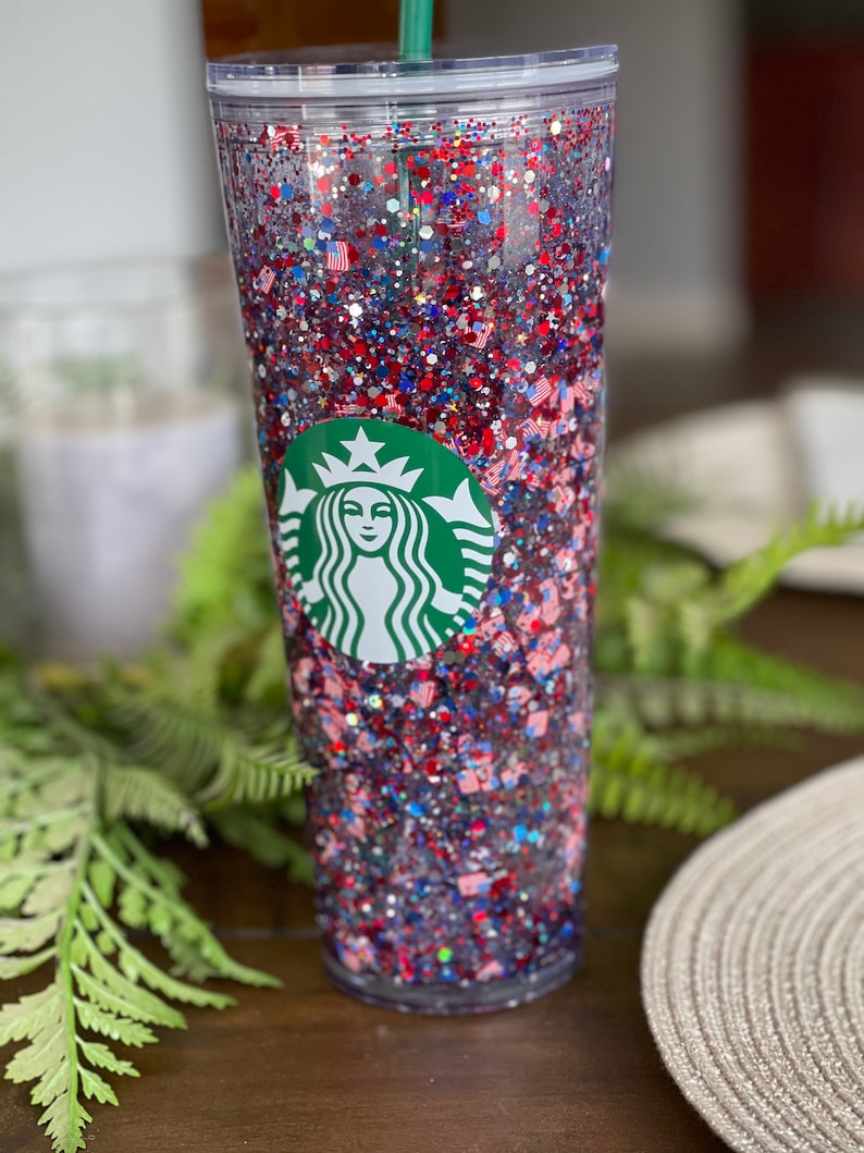 May include: A clear tumbler with a red, white and blue glitter design. The tumbler has the Starbucks logo on the front.