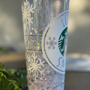 May include: A clear plastic tumbler with a white snowflake design and the text "Let it snow". The tumbler is filled with iridescent glitter.