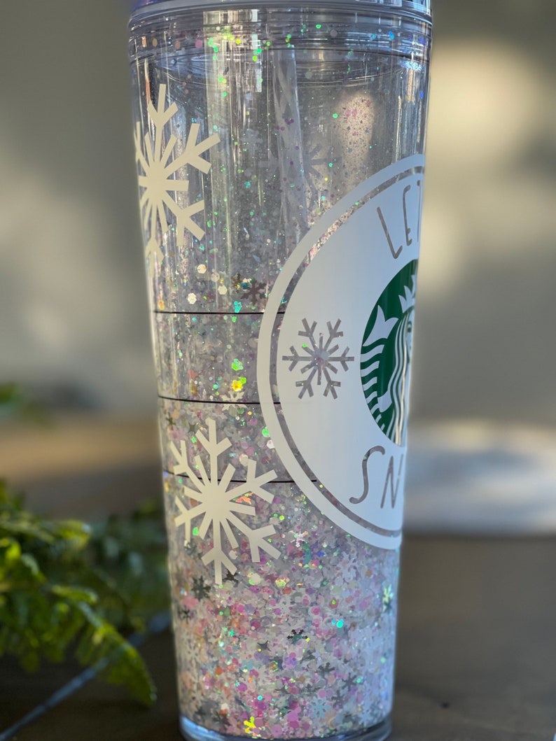 May include: A clear plastic tumbler with a white snowflake design and the text "Let it snow". The tumbler is filled with iridescent glitter.