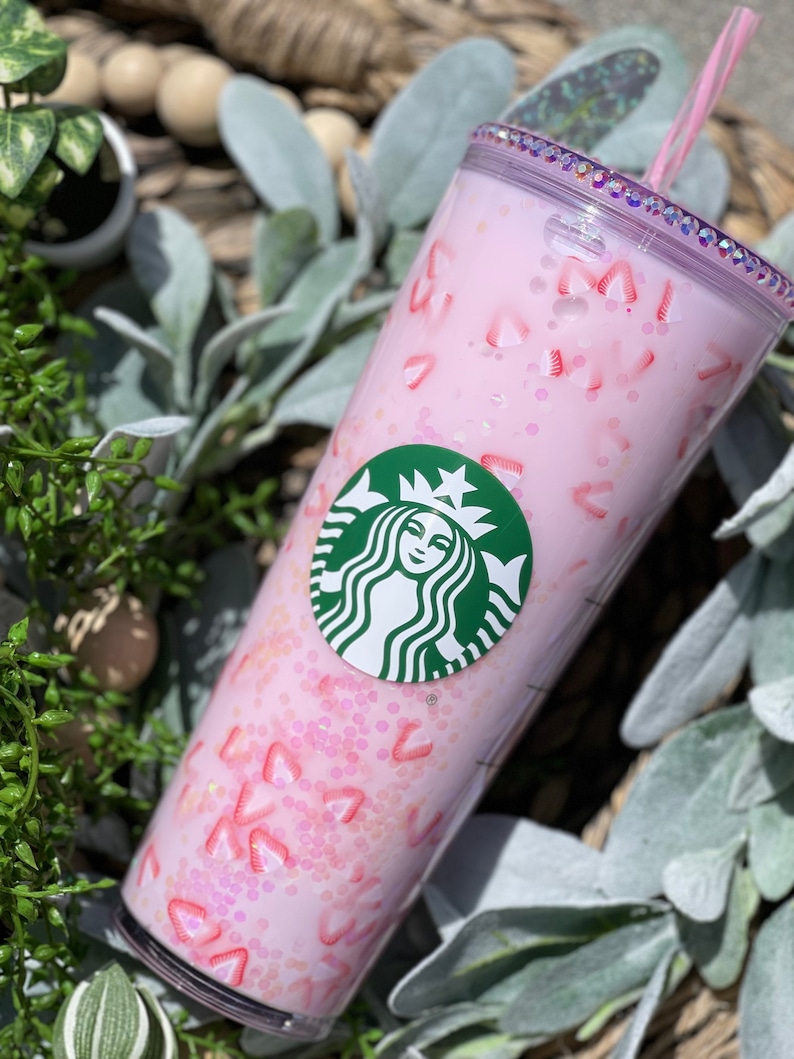 Peut inclure: Un gobelet rose avec un logo Starbucks et un motif de paillettes roses. Le gobelet a un couvercle transparent et une paille rose.