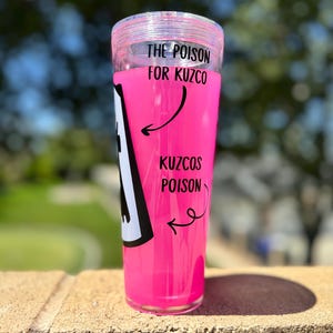 May include: A clear, cylindrical tumbler filled with bright pink liquid. The text "THE POISON FOR KUZCO" and "KUZCOS POISON" is printed on the side, along with a black and white graphic. The tumbler has a clear lid.