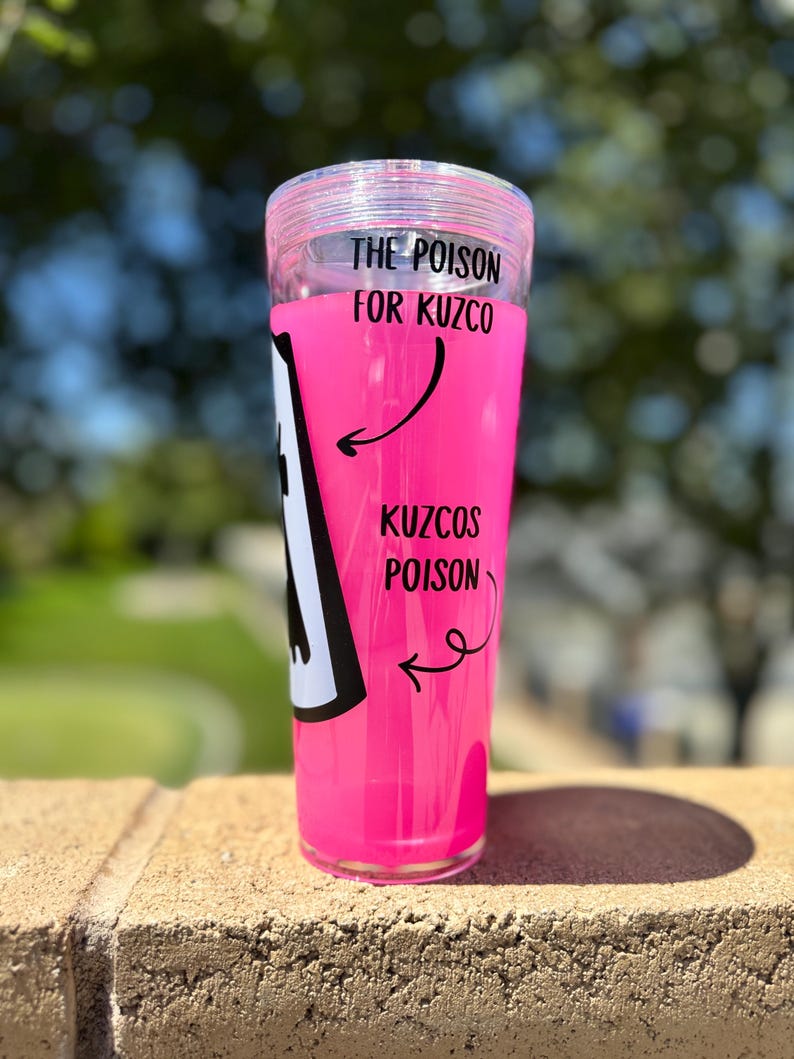 May include: A clear, cylindrical tumbler filled with bright pink liquid. The text "THE POISON FOR KUZCO" and "KUZCOS POISON" is printed on the side, along with a black and white graphic. The tumbler has a clear lid.