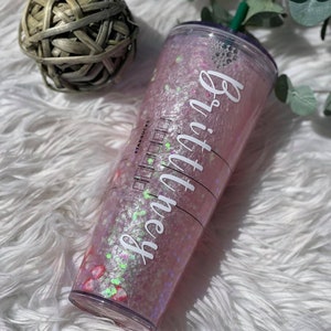May include: A clear plastic reusable cup with a pink and white glitter design. The cup has a green lid and a white sticker with the text "Brittney" on it.