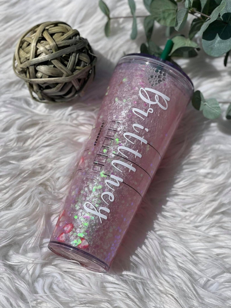 May include: A clear plastic reusable cup with a pink and white glitter design. The cup has a green lid and a white sticker with the text "Brittney" on it.