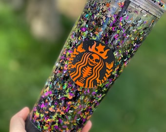 May include: A clear tumbler with a black lid and a black straw. The tumbler is filled with black, green, orange, and purple glitter. A black and orange Starbucks logo with a skeleton inside is on the side of the tumbler.