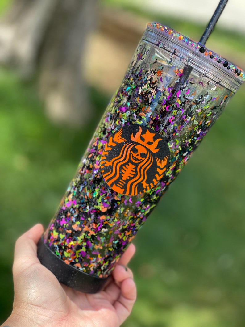 May include: A clear tumbler with a black lid and a black straw. The tumbler is filled with black, green, orange, and purple glitter. A black and orange Starbucks logo with a skeleton inside is on the side of the tumbler.