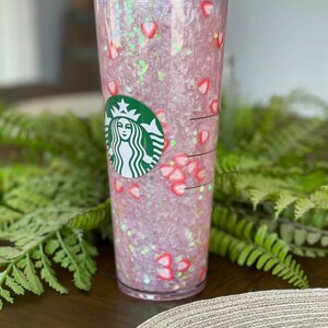 May include: A clear plastic tumbler with a pink and white glitter design and a green Starbucks logo. The tumbler is filled with pink and white glitter and has a straw.