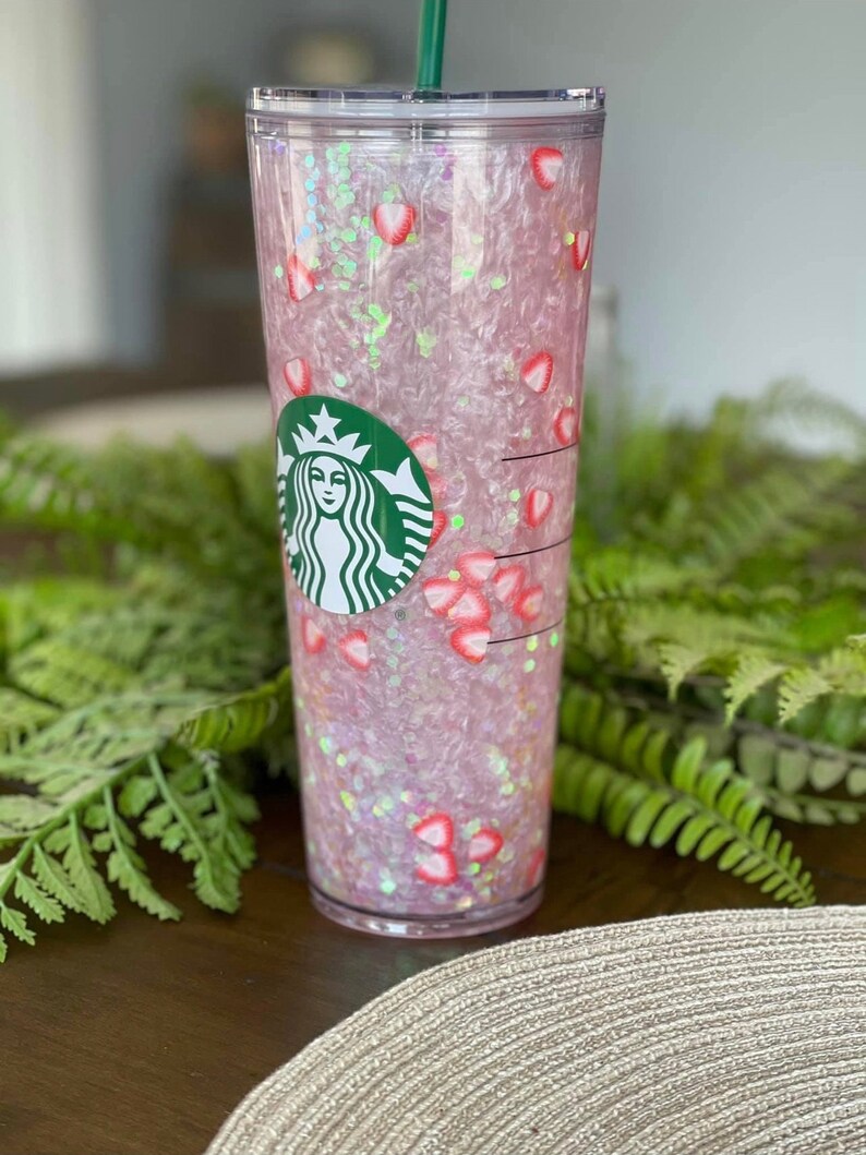 May include: A clear plastic tumbler with a pink and white glitter design and a green Starbucks logo. The tumbler is filled with pink and white glitter and has a straw.