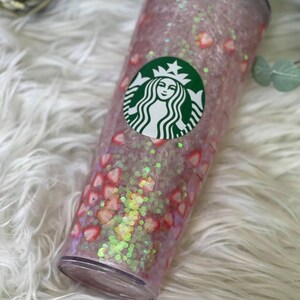 May include: A clear plastic tumbler with a pink and white glitter base and a green Starbucks logo. The tumbler is decorated with pink and white hearts and glitter.