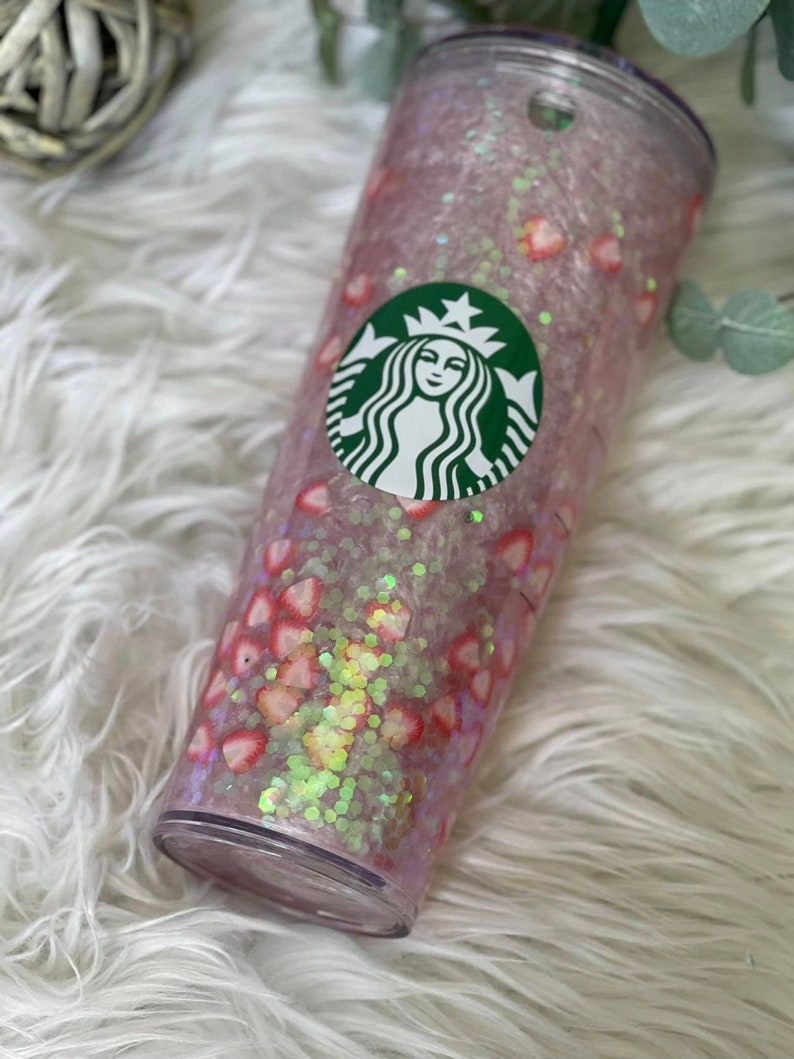 May include: A clear plastic tumbler with a pink and white glitter base and a green Starbucks logo. The tumbler is decorated with pink and white hearts and glitter.