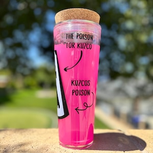 May include: A clear glass tumbler with a cork lid, filled with bright pink liquid. The text "THE POISON FOR KUZCO" is printed on the top, with "KUZCOS POISON" and an arrow on the side. The tumbler is sitting on a brick surface.